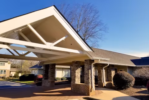 Exterior view of a senior living facility entrance with a covered driveway supported by stone pillars, surrounded by bushes and trees under a clear blue sky.