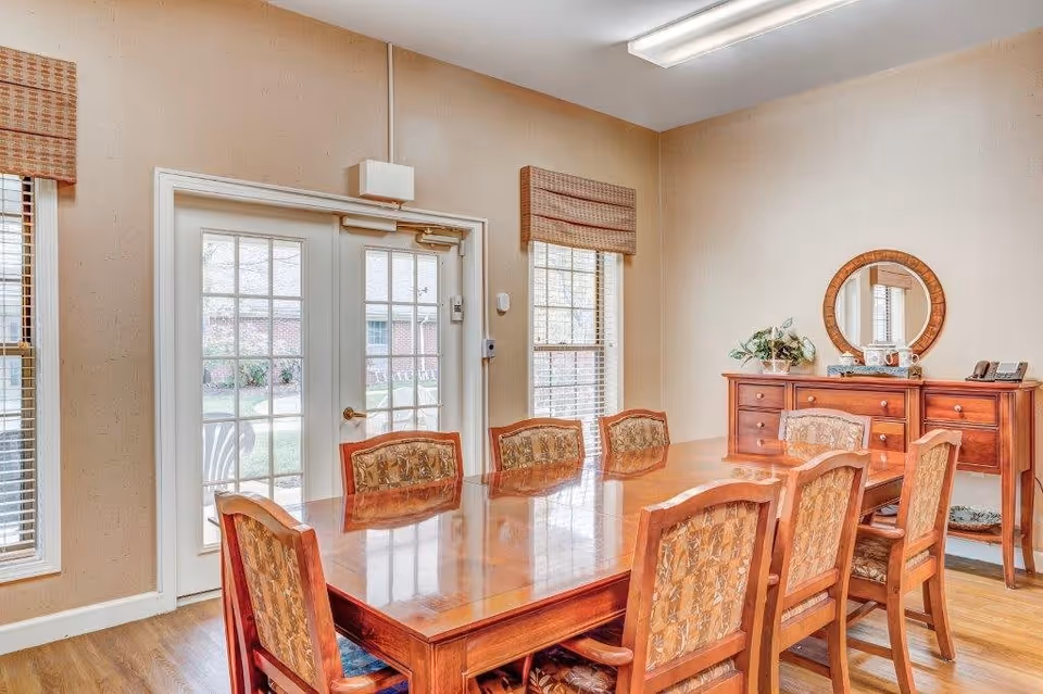 A dining room with a large wooden table surrounded by eight upholstered chairs. The room has beige walls, two windows with blinds, and a set of French doors leading outside. Against one wall is a wooden sideboard with a round mirror above it and some decorative items on top.