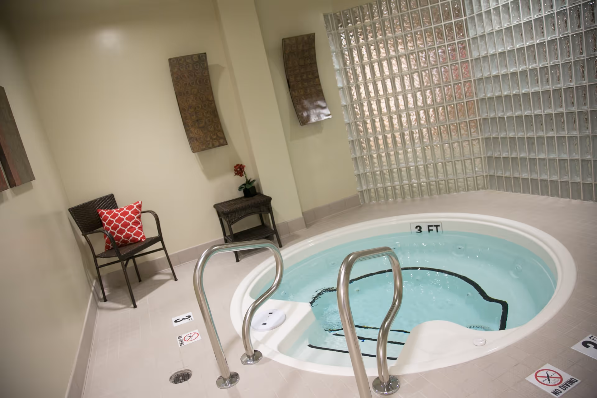 Indoor hot tub with clear water and stainless steel handrails, surrounded by beige tiled floor and walls. Two wicker chairs with a red patterned cushion and a small table with a potted plant are placed against the wall. The background features a glass block wall.