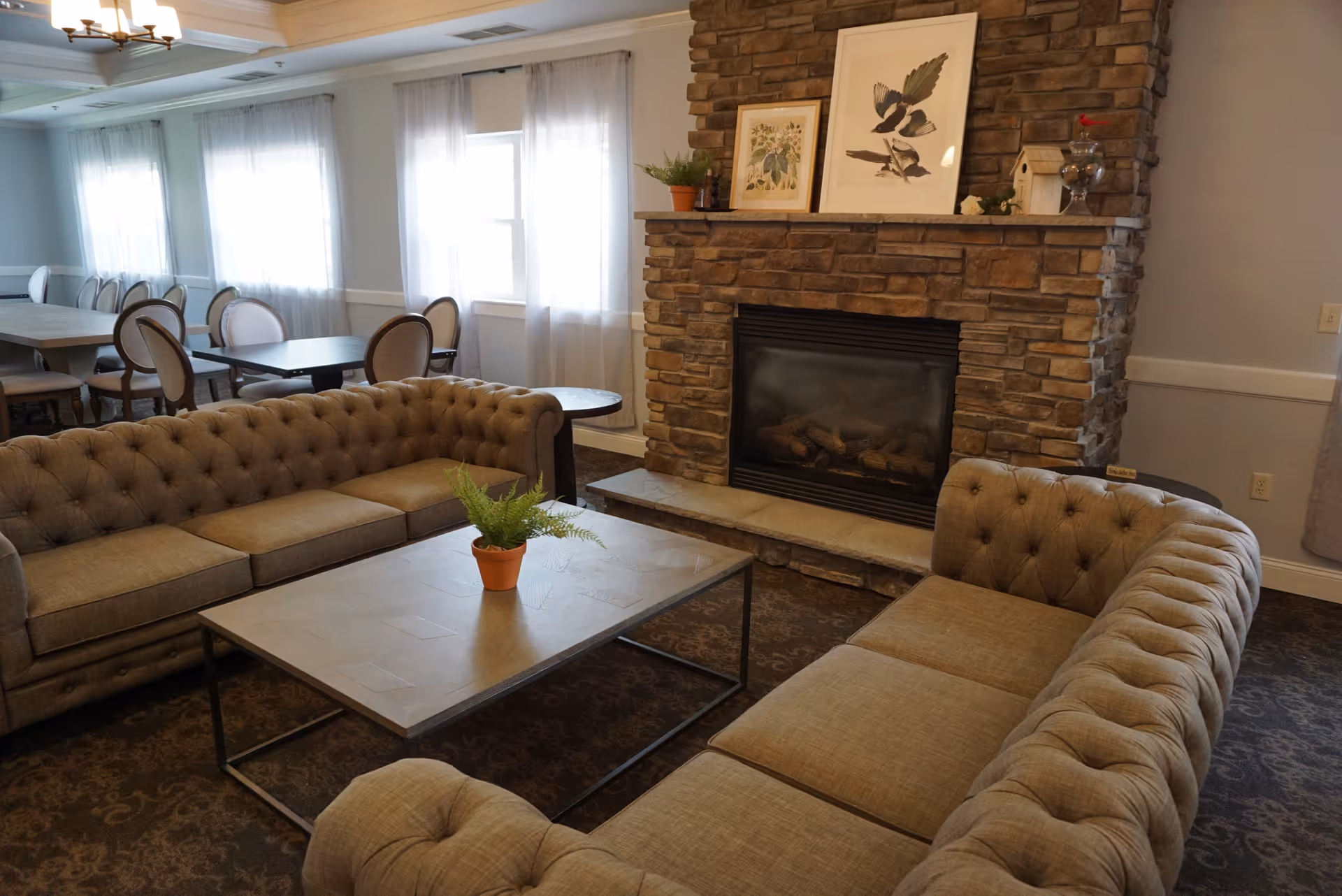 A cozy living room area with two tufted beige sofas arranged around a rectangular coffee table with a small potted plant. In the background, there is a stone fireplace with framed artwork and decorative items on the mantel. Behind the seating area, there are dining tables and chairs near windows with sheer white curtains.