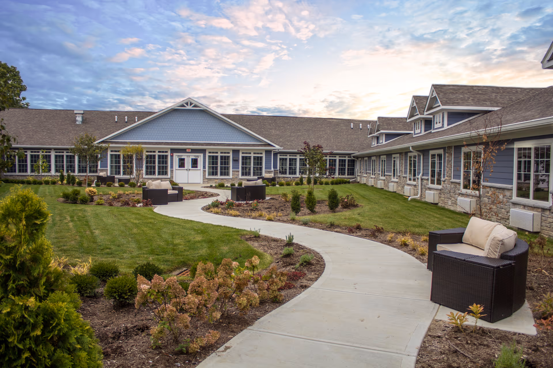 Landscaped courtyard with a curved concrete walkway, patio seating, and a single-story senior living building in the background under a partly cloudy sky.