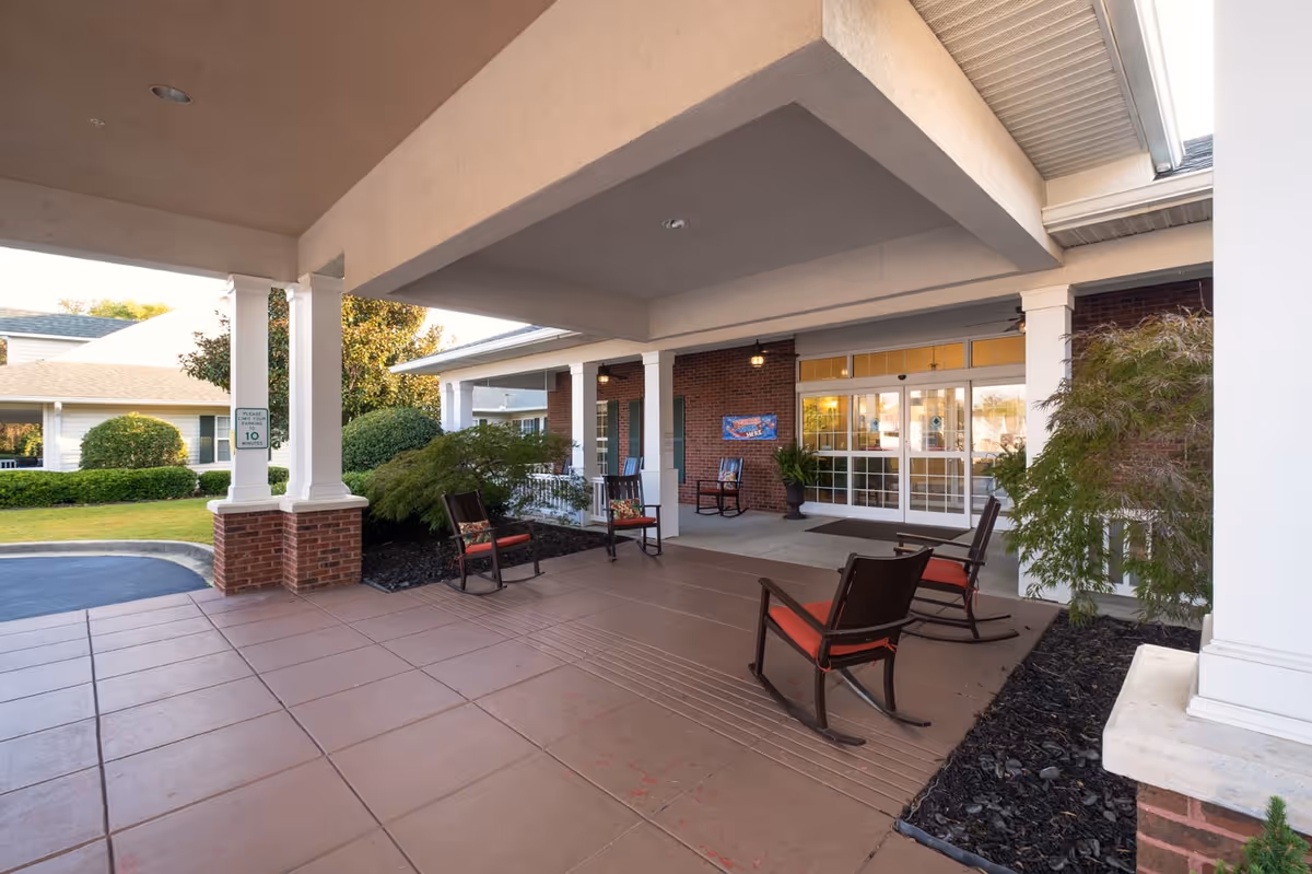 Covered entrance area of The Pines at Greenville Assisted Living & Memory Care with several rocking chairs featuring red cushions, brick pillars, and glass double doors leading inside. There are shrubs and greenery around the entrance and a sign indicating a 10-minute parking limit.