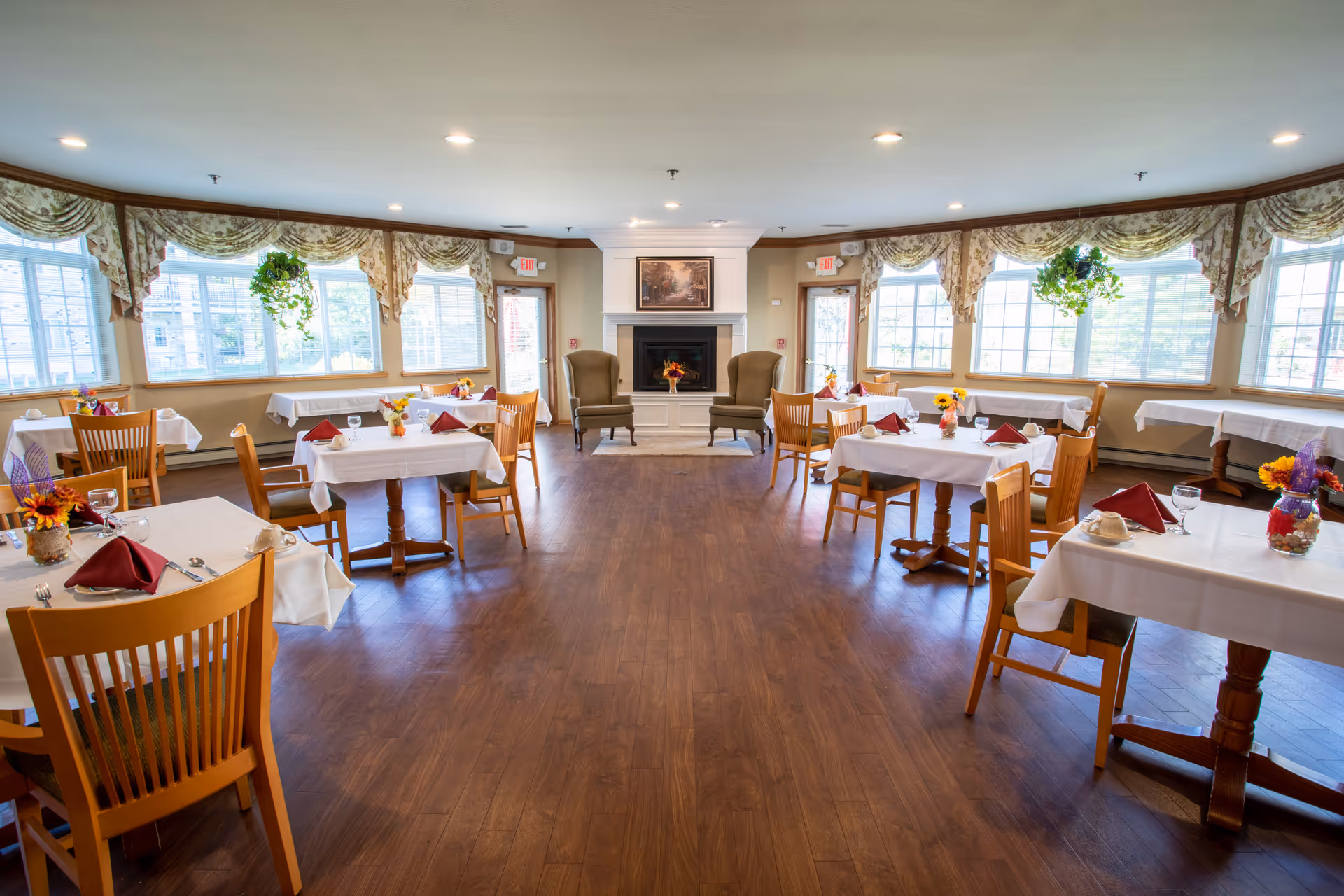 A bright dining room with wooden floors and multiple tables covered with white tablecloths, each set with red napkins, cups, and glasses. The room has large windows with floral valances and hanging plants. At the far end, there is a white fireplace flanked by two armchairs and a framed picture above it.