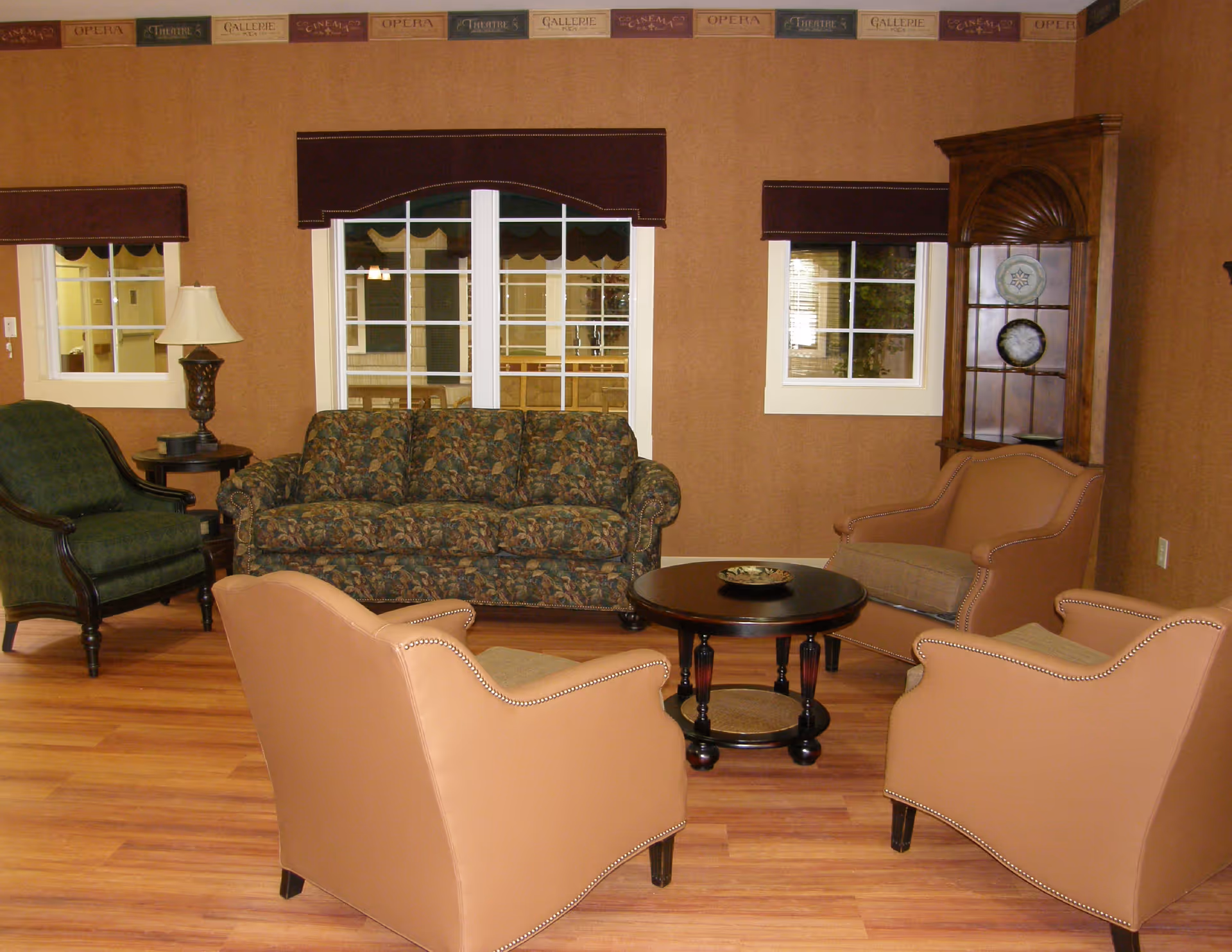 A cozy living room with a floral patterned sofa, two tan armchairs, a green armchair, a round wooden coffee table, a side table with a lamp, and a wooden corner cabinet displaying decorative plates. The walls are brown with a decorative border near the ceiling, and there are three windows with dark valances.