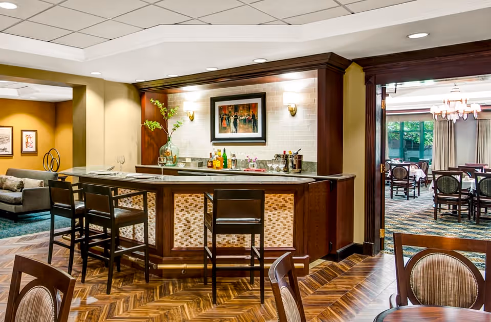 Interior view of a senior living facility lounge area featuring a wooden bar with high chairs, bottles, and glassware on the counter. The bar is decorated with a large vase with green branches and a framed painting on the wall behind it. To the right, there is an adjoining dining room with tables and chairs, large windows, and curtains. The floor has a wood pattern, and the ceiling has recessed lighting.