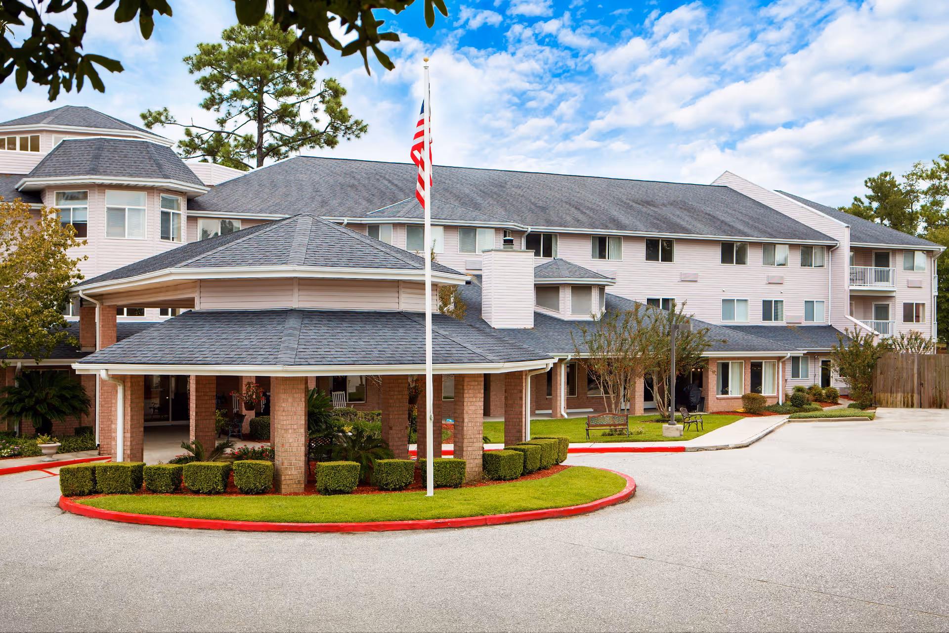 Front exterior of a multi-story senior living building with a circular porte-cochere, an American flagpole, and landscaped driveway.