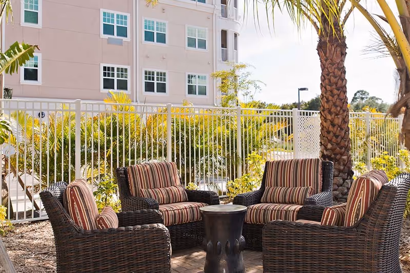 Outdoor patio with four wicker armchairs with striped cushions arranged around a small table beside a palm tree and a fenced apartment building.