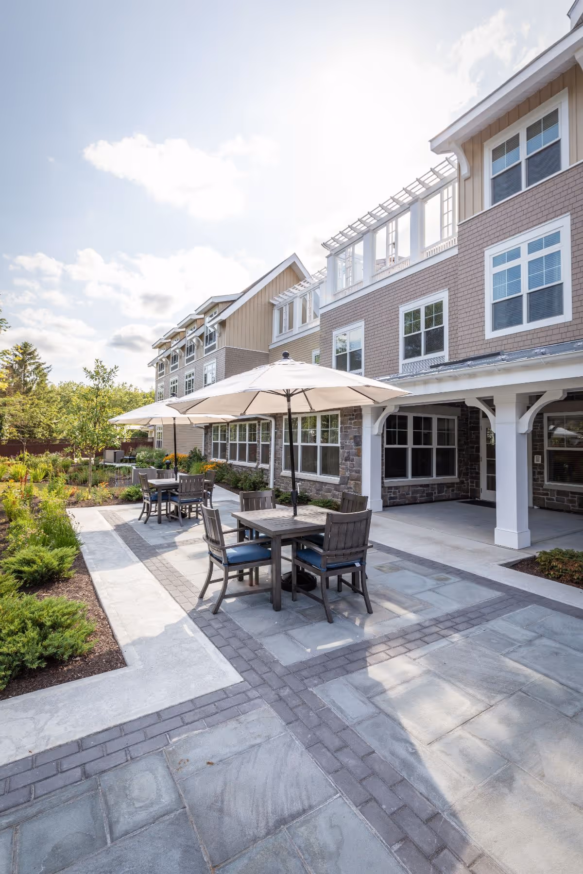 Outdoor patio area at Sunrise of Wilton with several tables and chairs under large white umbrellas, surrounded by landscaped greenery and adjacent to a multi-story building with large windows.