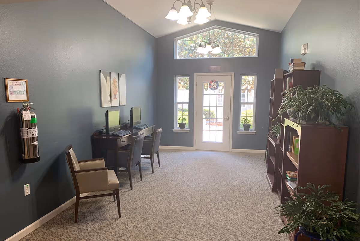 A quiet interior room with two computer desks and chairs along the left wall, a fire extinguisher mounted on the wall, and bookshelves with plants and books on the right. At the far end, there is a glass door with windows on either side and a transom window above, letting in natural light.