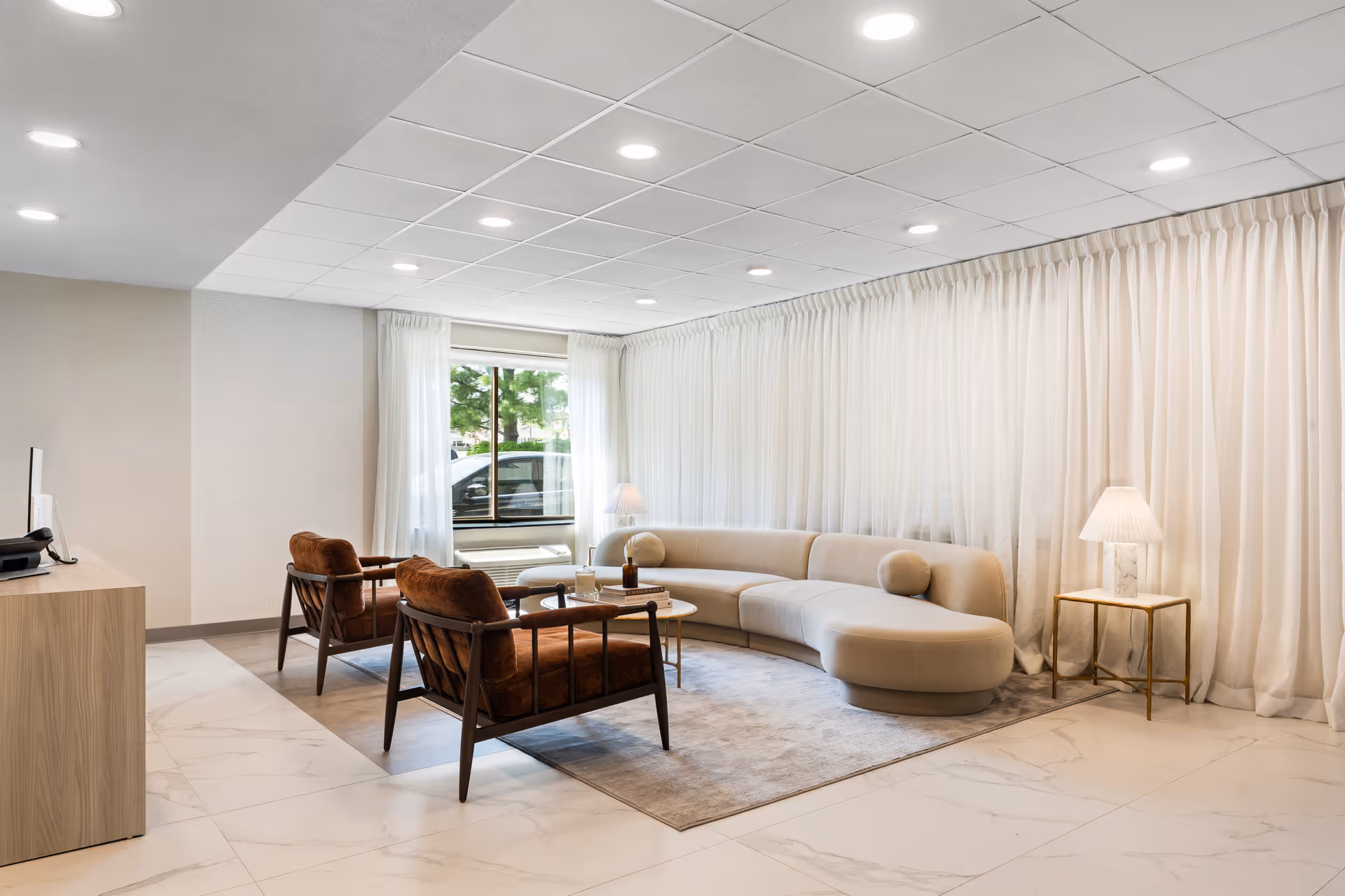 A modern and bright living room area with a curved beige sofa, two brown cushioned armchairs, a small round coffee table, and a side table with a lamp. The room has large windows with sheer white curtains, a light-colored tiled floor, and recessed ceiling lights.