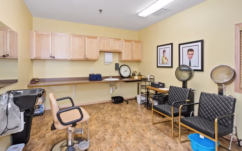 Interior of a salon room with two black salon chairs with hair dryers, a beige salon chair in front of a black sink, wooden cabinets mounted on a yellow wall, a large clock on the counter, and two framed portraits on the wall.