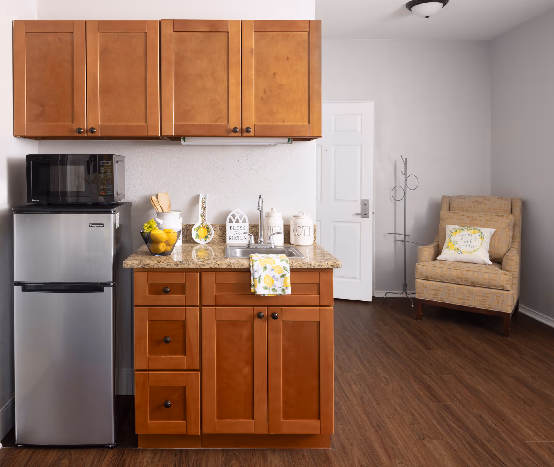 A small kitchen area with wooden cabinets, a granite countertop with a sink, and a stainless steel mini refrigerator topped with a microwave. On the counter are decorative items including a bowl of lemons, a utensil holder, and canisters labeled sugar and coffee. To the right, there is a beige armchair with a decorative pillow and a coat rack near a white door. The floor is wood and the walls are light-colored.