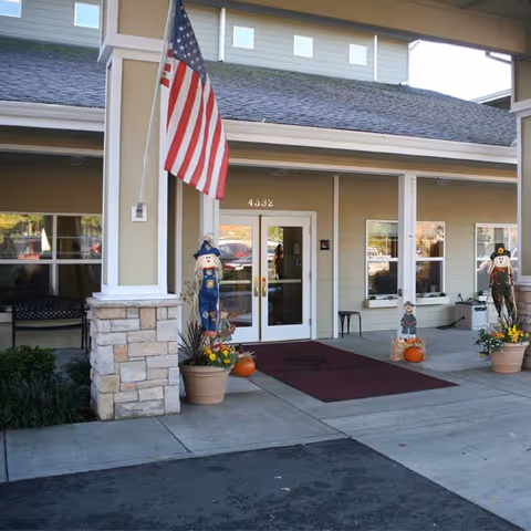 Entrance of a senior living facility with double glass doors, an American flag mounted on a pillar, and seasonal decorations including scarecrows and pumpkins in pots on either side of the entrance. The building exterior is beige with stone accents and several windows.