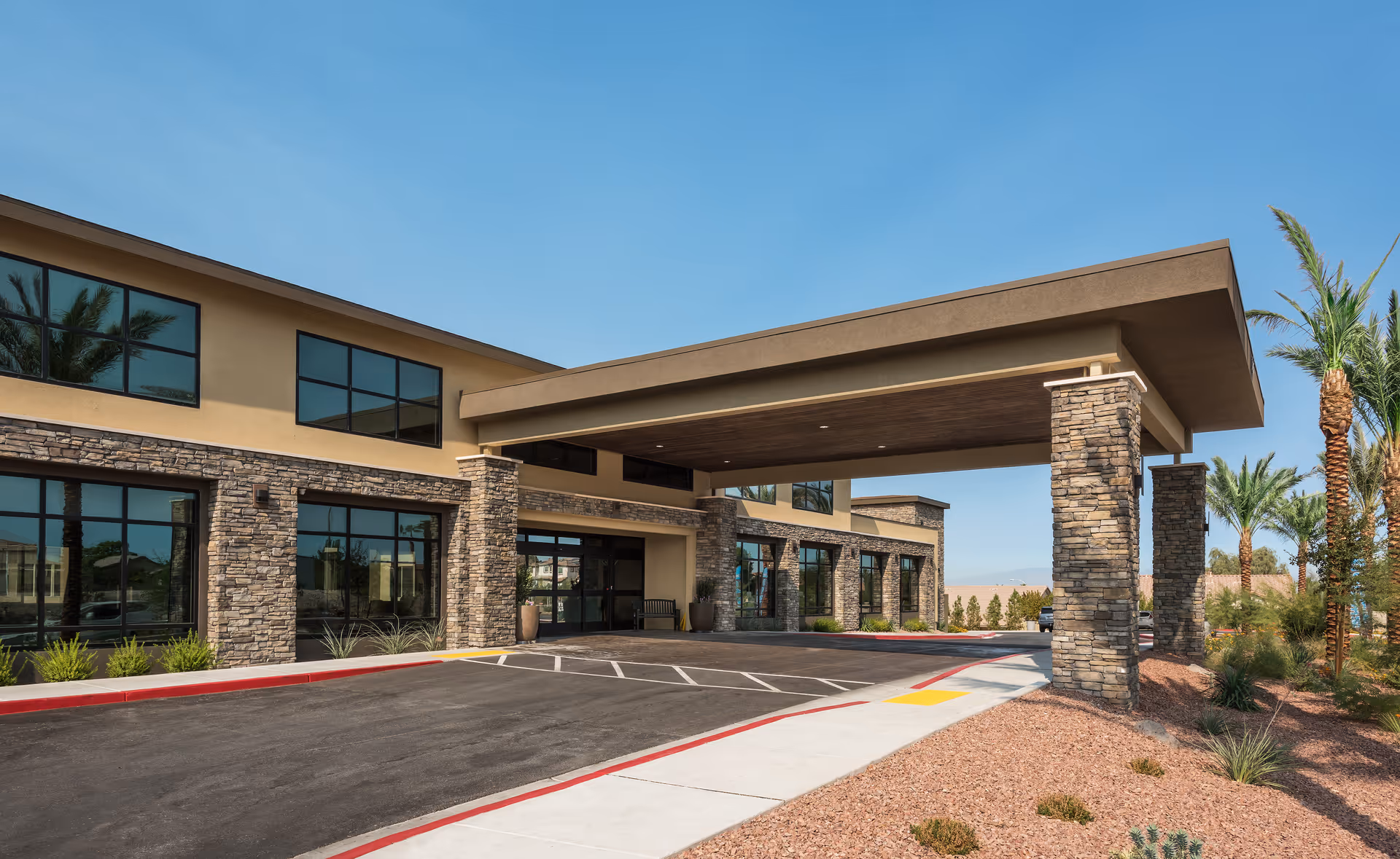 Exterior view of Carnegie Heights at Henderson showing the building entrance with a covered drop-off area supported by stone pillars, large windows, and desert landscaping with palm trees under a clear blue sky.