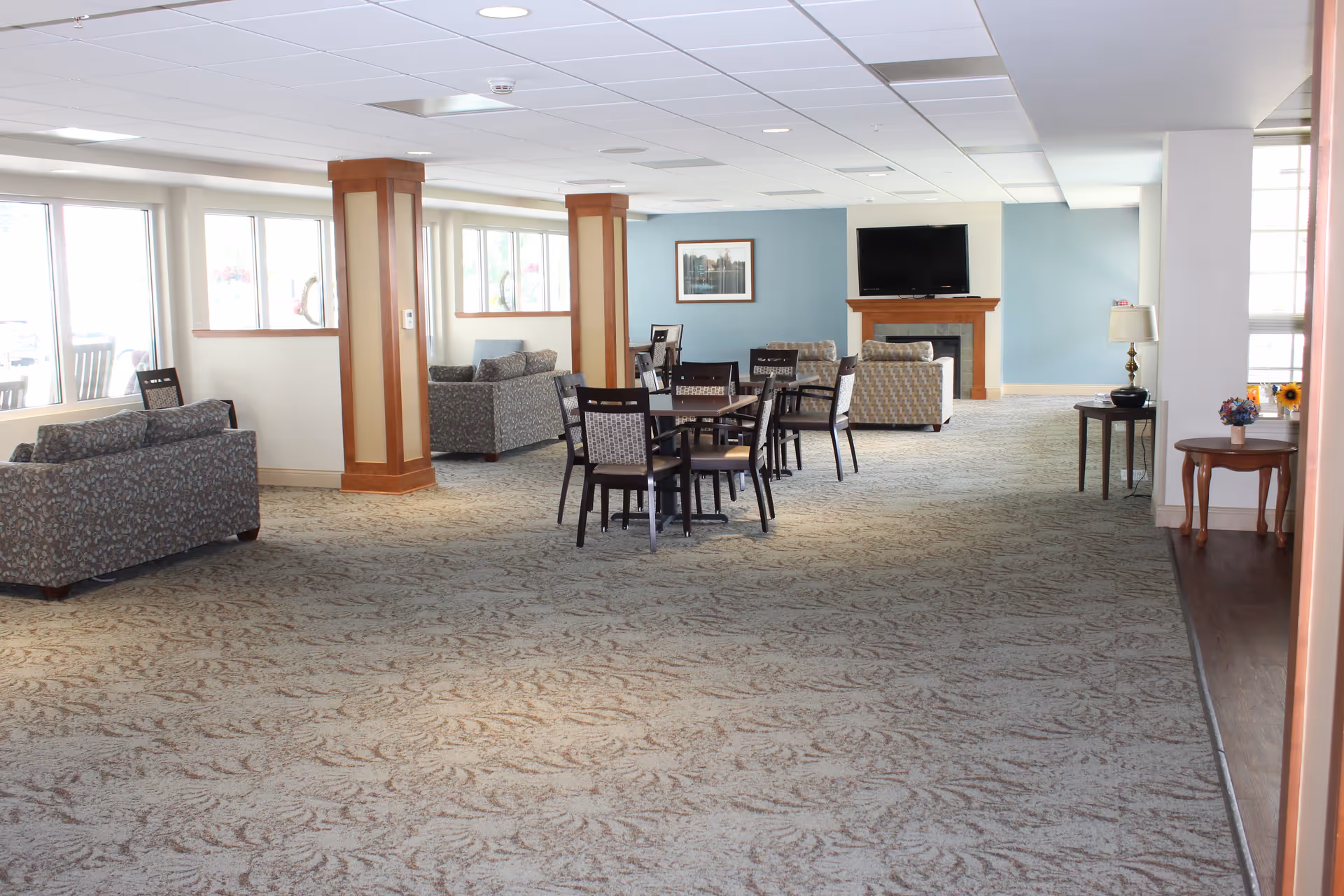 A spacious senior living common area with patterned carpet, several seating arrangements including sofas and chairs, a few tables with chairs, large windows letting in natural light, a wall-mounted TV above a fireplace, and decorative columns.