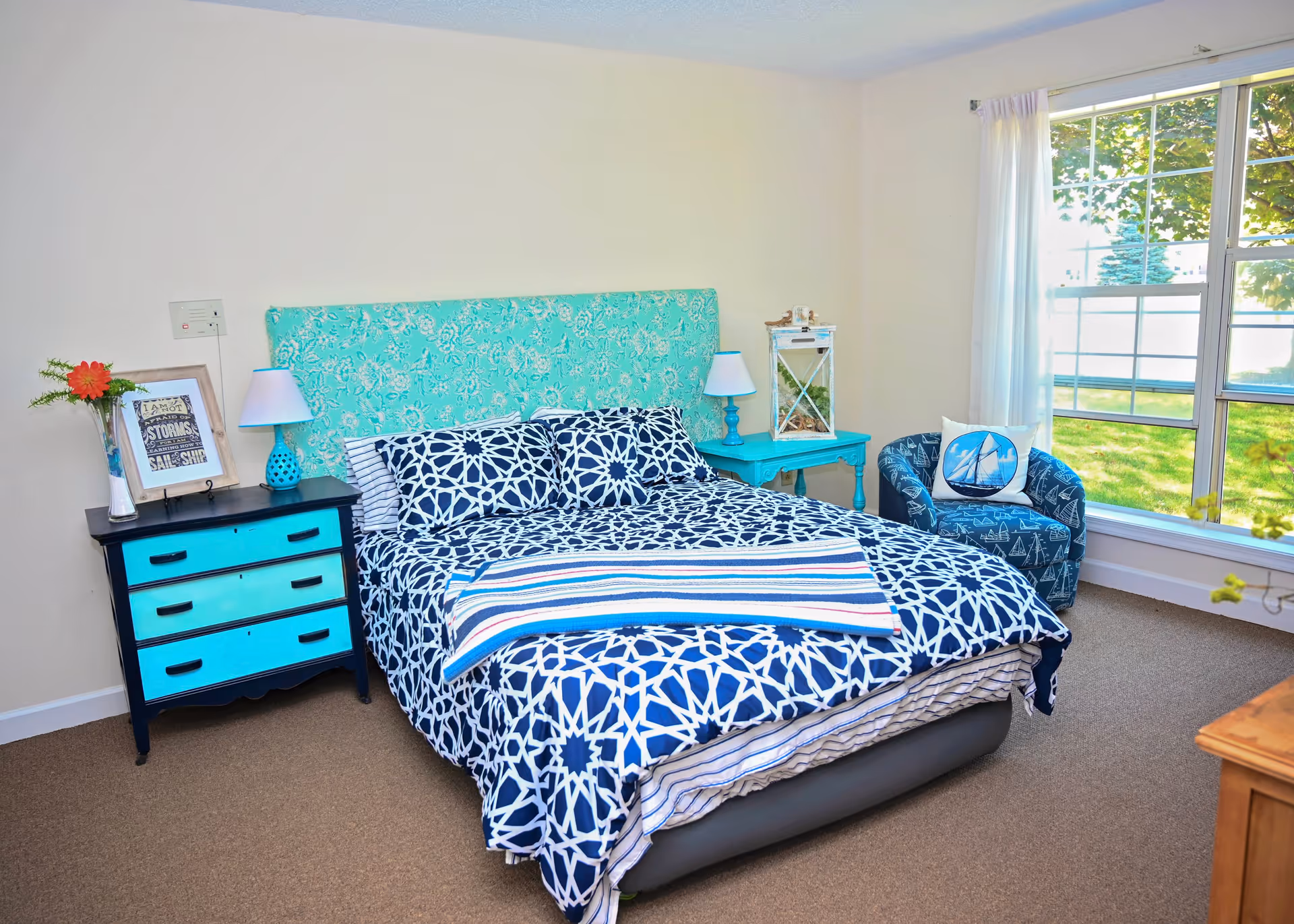 A bright bedroom with a bed featuring a turquoise patterned headboard and blue and white geometric bedding. There is a small black and turquoise dresser with a lamp and framed picture on it to the left of the bed. To the right of the bed is a turquoise side table with a white lantern on top. A blue armchair with a sailboat pillow is positioned near a large window with white curtains, letting in natural light and showing green grass and trees outside.