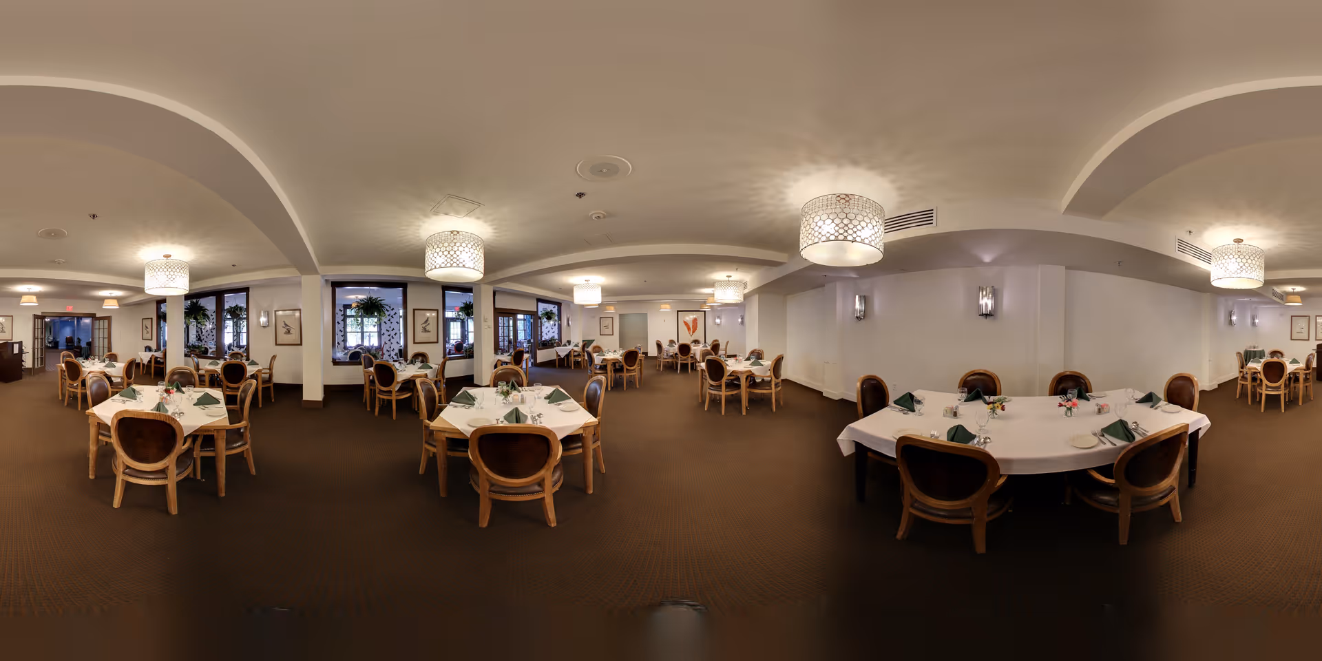Dining room with round tables set with white tablecloths, chairs, place settings, and hanging pendant lights.