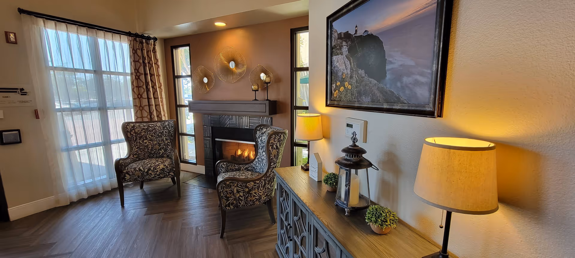 A cozy seating area in a senior living facility featuring two patterned armchairs positioned near a lit fireplace. The room has large windows with sheer and patterned curtains allowing natural light to enter. A wooden sideboard with decorative items, including a lantern, small plants, and two table lamps, is placed against the wall. Above the sideboard hangs a framed picture of a lighthouse on a rocky coast.