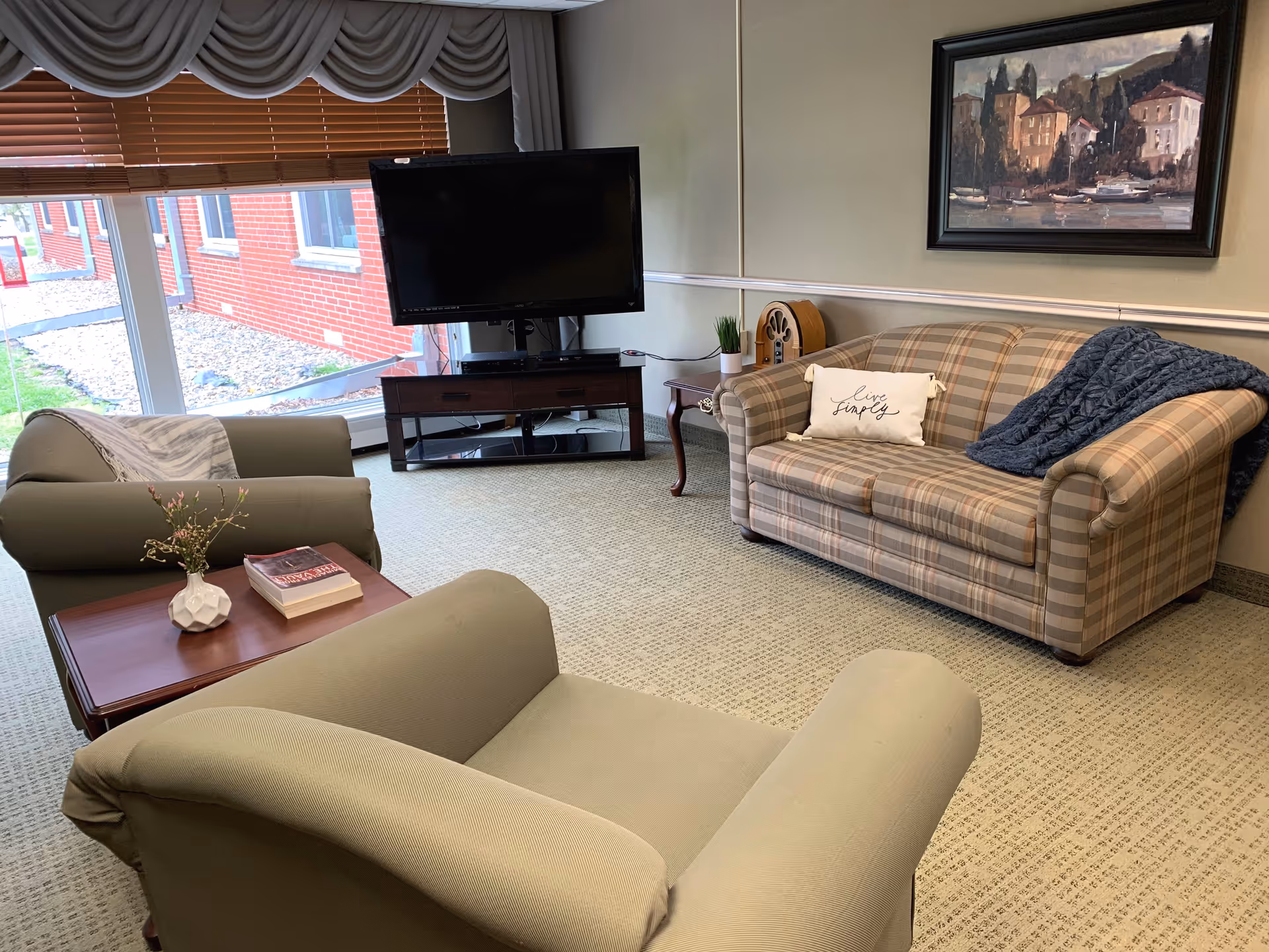 A cozy living room with a plaid sofa adorned with a 'Live Simply' pillow and a dark blue throw blanket. Two beige armchairs face a wooden coffee table with a small vase of flowers and a book. A flat-screen TV sits on a dark wooden stand near large windows with wooden blinds and a scalloped valance. A framed painting hangs on the wall above the sofa.