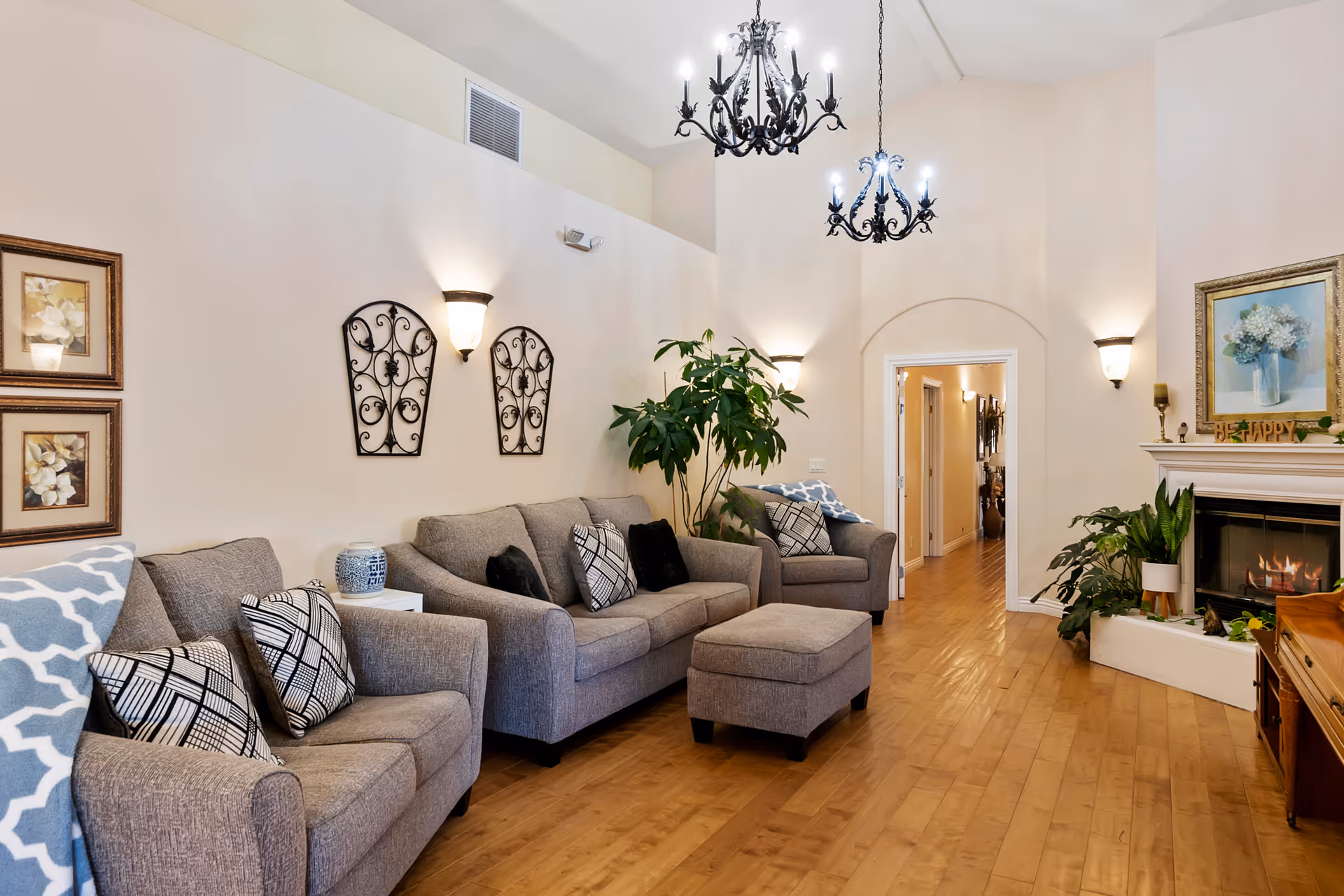 Bright living room with gray sofas and ottoman, patterned pillows, chandeliers, a fireplace, plants, and hardwood floors.