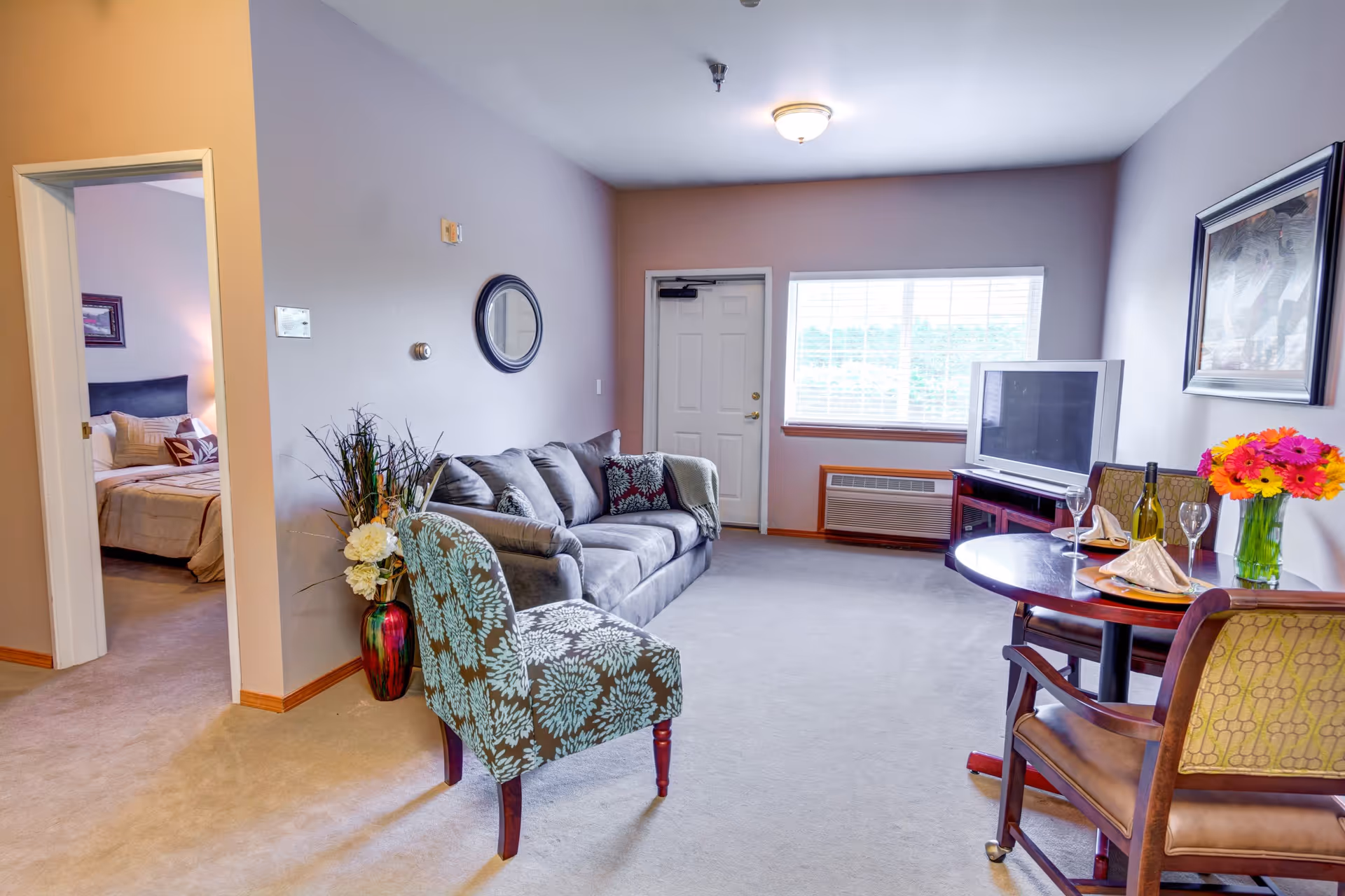 Furnished living room with a gray sofa, patterned armchair, small dining table set, television, and a doorway leading to a bedroom.