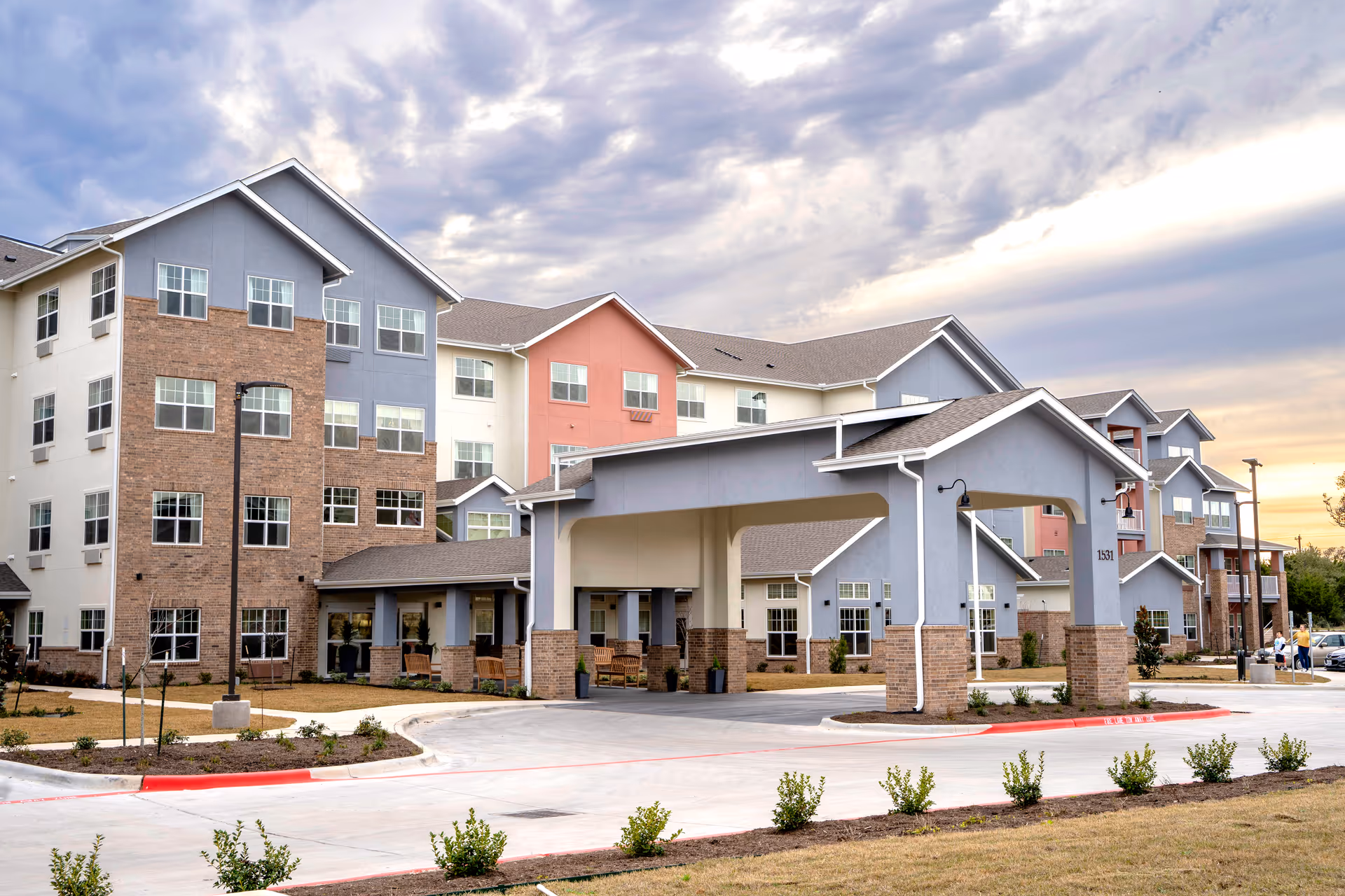 Exterior view of a multi-story senior living facility with a covered entrance driveway, landscaped grounds, and a partly cloudy sky at sunset.
