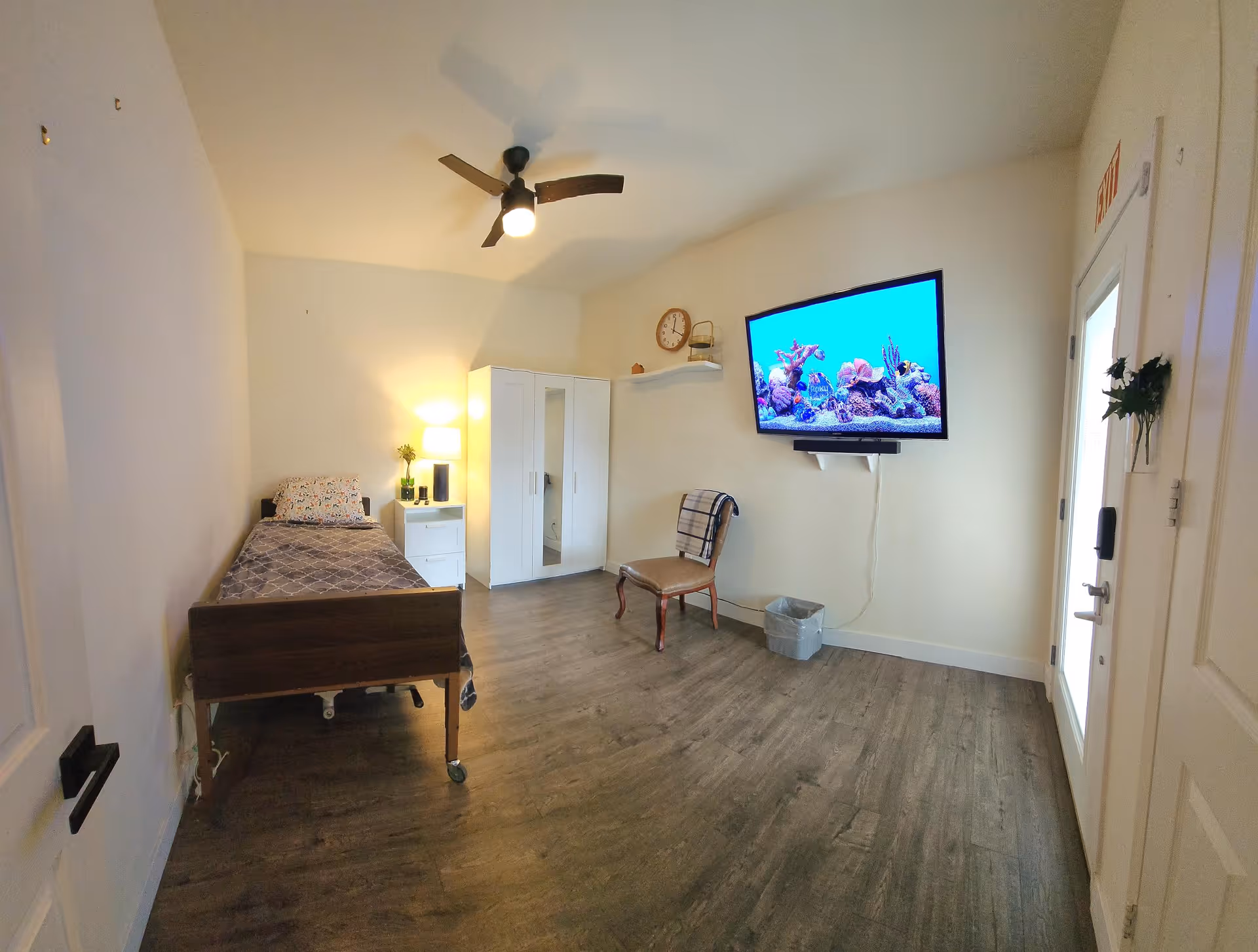 A simple, clean bedroom in an assisted living facility featuring a single bed with patterned bedding, a white nightstand with a lamp and plant, a white wardrobe, a wooden chair with a plaid blanket draped over it, a wall-mounted TV displaying an aquarium scene, a clock on a small wall shelf, and a door with an exit sign above it.