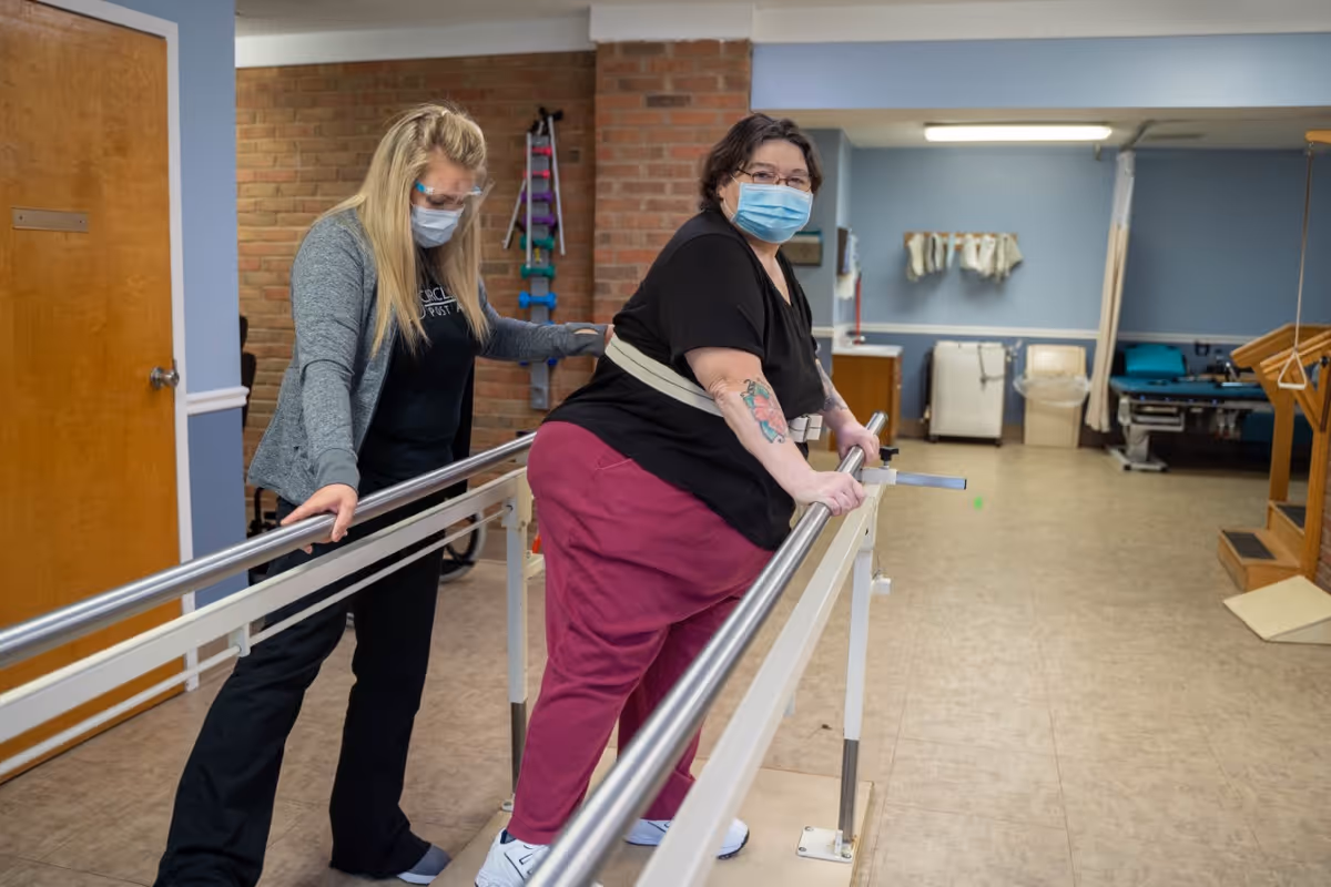 A woman wearing a mask and maroon pants is using parallel bars for physical therapy in a rehabilitation room, assisted by a masked therapist in black scrubs and a gray cardigan. The room has a brick wall, exercise equipment, and medical supplies in the background.