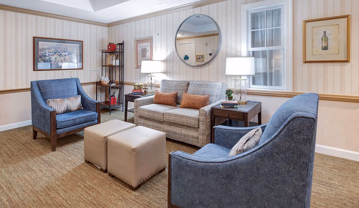 A cozy living room with two blue armchairs, a beige loveseat with two brown cushions, two beige ottomans, a round mirror on the wall, two table lamps on dark wooden side tables, framed artwork, and a small shelving unit in the corner.