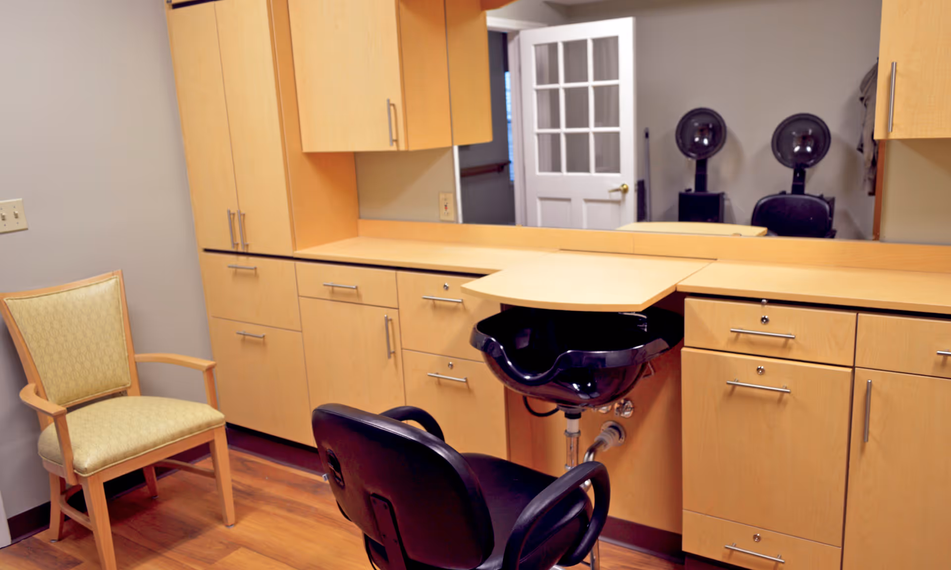 Interior view of a hair salon area in a senior living facility with a black salon chair facing a black hair washing basin. Light wood cabinets and drawers line the wall beneath a large mirror. A beige upholstered chair is positioned to the left, and two black hair dryer chairs are visible in the mirror's reflection. The room has wood flooring and a white door with glass panels is open in the background.