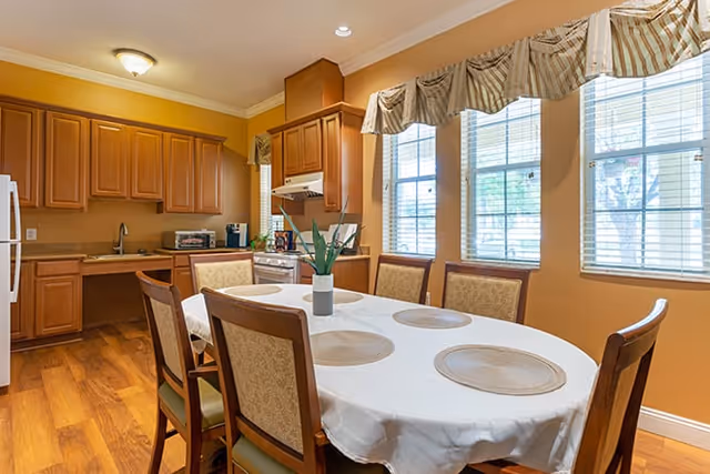 A dining area with a rectangular table covered with a white tablecloth and six chairs around it. The table has six round placemats and a small plant centerpiece. The room has large windows with striped valances allowing natural light to enter. In the background, there is a kitchen with wooden cabinets, a white refrigerator, a stove, and a countertop with small appliances.
