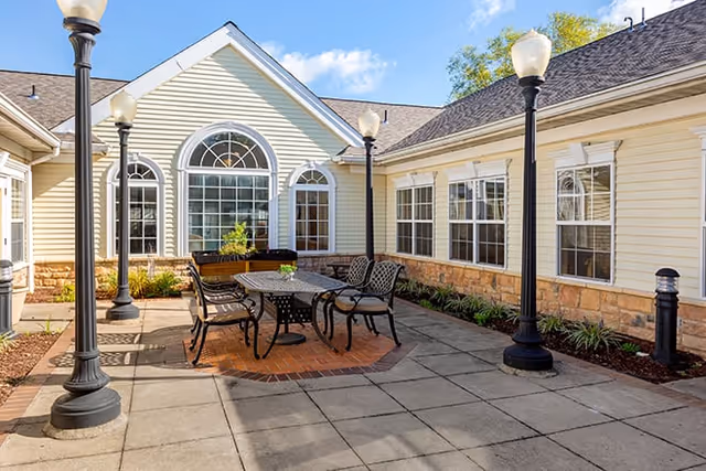Outdoor courtyard area at Brookdale Westampton with a metal table and chairs set on a brick and concrete patio, surrounded by cream-colored building walls with large windows and black lamp posts.