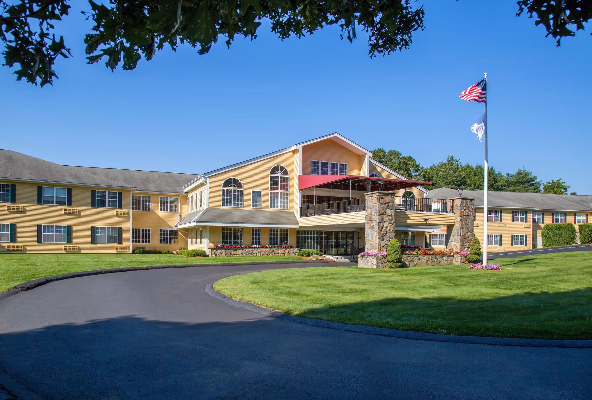 Exterior view of a large, yellow senior living facility building with multiple windows and a gray roof. The building has a covered entrance supported by stone pillars and a red awning above a balcony. A curved driveway leads to the entrance, and a flagpole with the American flag and another flag stands on a well-maintained green lawn. Trees and clear blue sky are visible in the background.