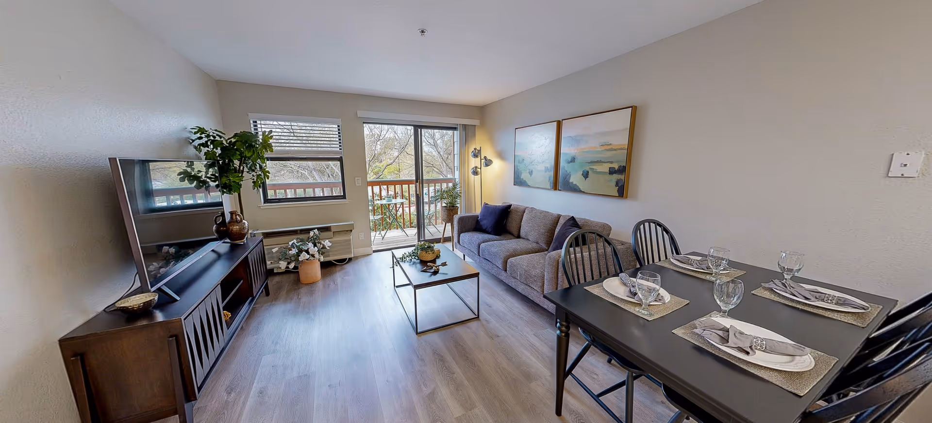 A bright and modern living room in a senior living facility with a gray sofa, two framed landscape paintings on the wall, a coffee table with a plant, a TV on a wooden stand, and a dining table set for four with plates, napkins, and glasses. Large windows and a sliding glass door lead to a balcony with outdoor seating and a view of trees.