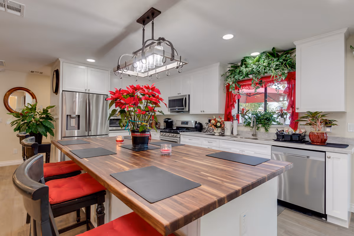A modern kitchen with a large wooden island countertop surrounded by black chairs with red cushions. The kitchen features white cabinets, stainless steel appliances including a refrigerator, microwave, and dishwasher. There are several potted plants and flowers on the countertop and windowsill, with a window adorned with red curtains. A rectangular chandelier with multiple lights hangs above the island.