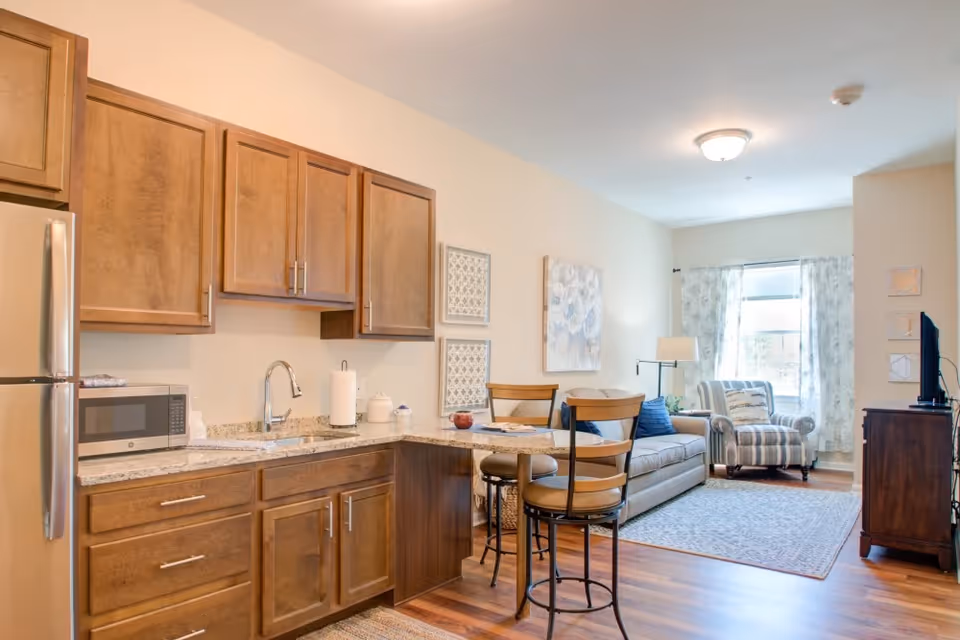 A bright and cozy living space featuring a small kitchen area with wooden cabinets, a granite countertop, a microwave, and a stainless steel refrigerator. Adjacent to the kitchen is a dining area with two high chairs at a counter. The living room includes a beige sofa with blue cushions, a striped armchair, a floor lamp, a window with sheer curtains, and a TV on a wooden stand. The room has wooden flooring and light-colored walls with decorative wall art.