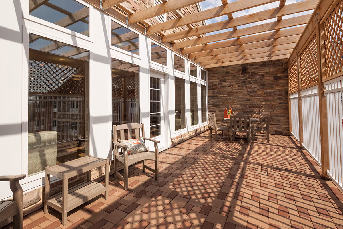A sunlit outdoor patio area with a wooden pergola overhead casting shadows on the brick floor. The patio features wooden chairs, a small table with a decorative pillow on one chair, and a dining table with chairs against a stone wall. Large windows and a door lead inside the building.