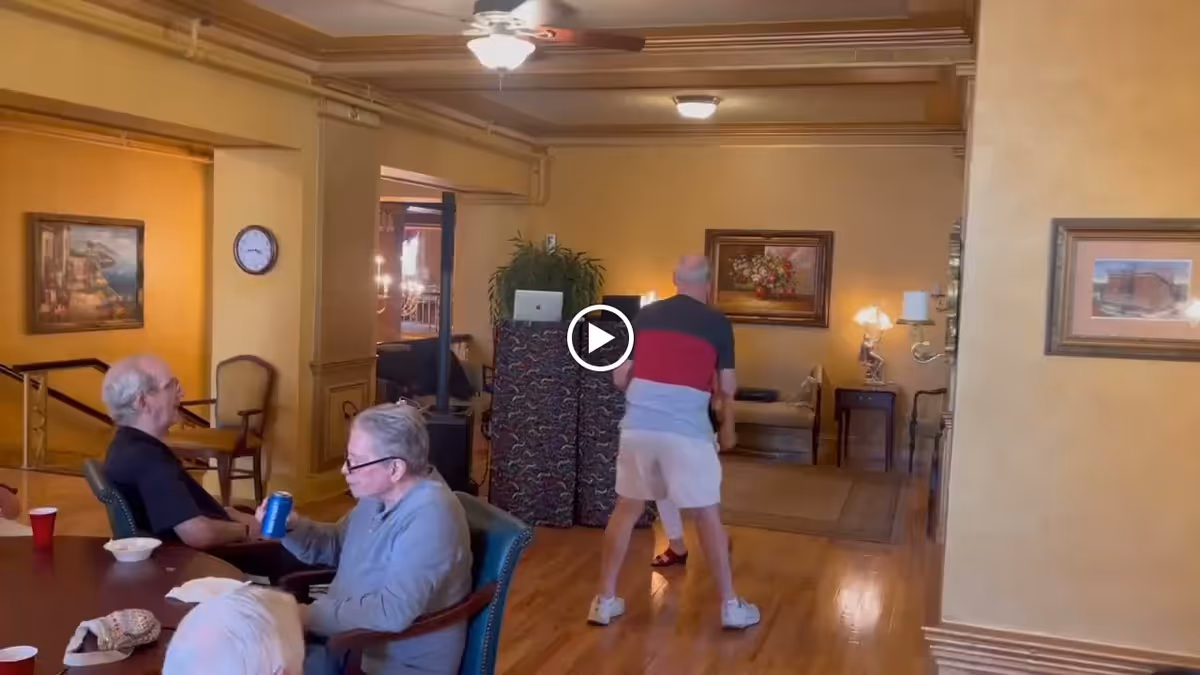 A common area in a senior living facility with several elderly people. Two men are seated at a table, one holding a can of soda, while another person stands in the background near a decorative divider. The room has warm yellow walls, framed paintings, wooden furniture, and a ceiling fan with a light.