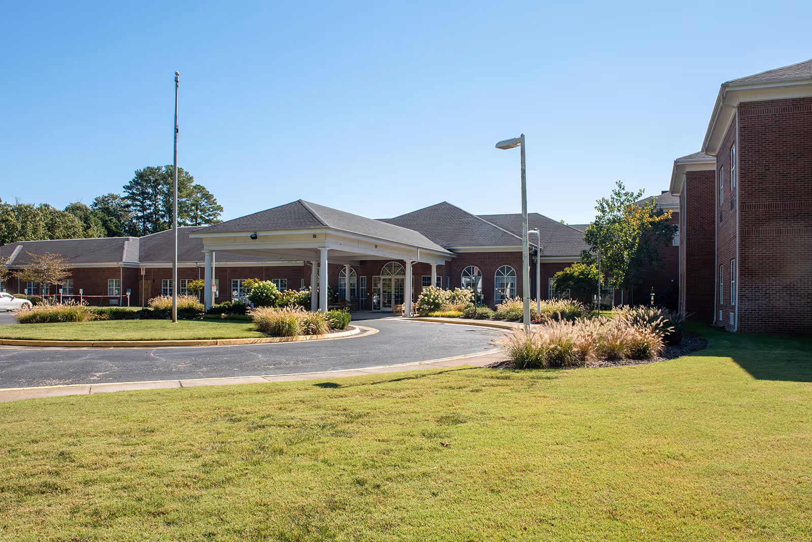 Front exterior view of a senior living facility building with a covered entrance, brick walls, arched windows, a driveway, landscaped bushes, and a grassy lawn under a clear blue sky.