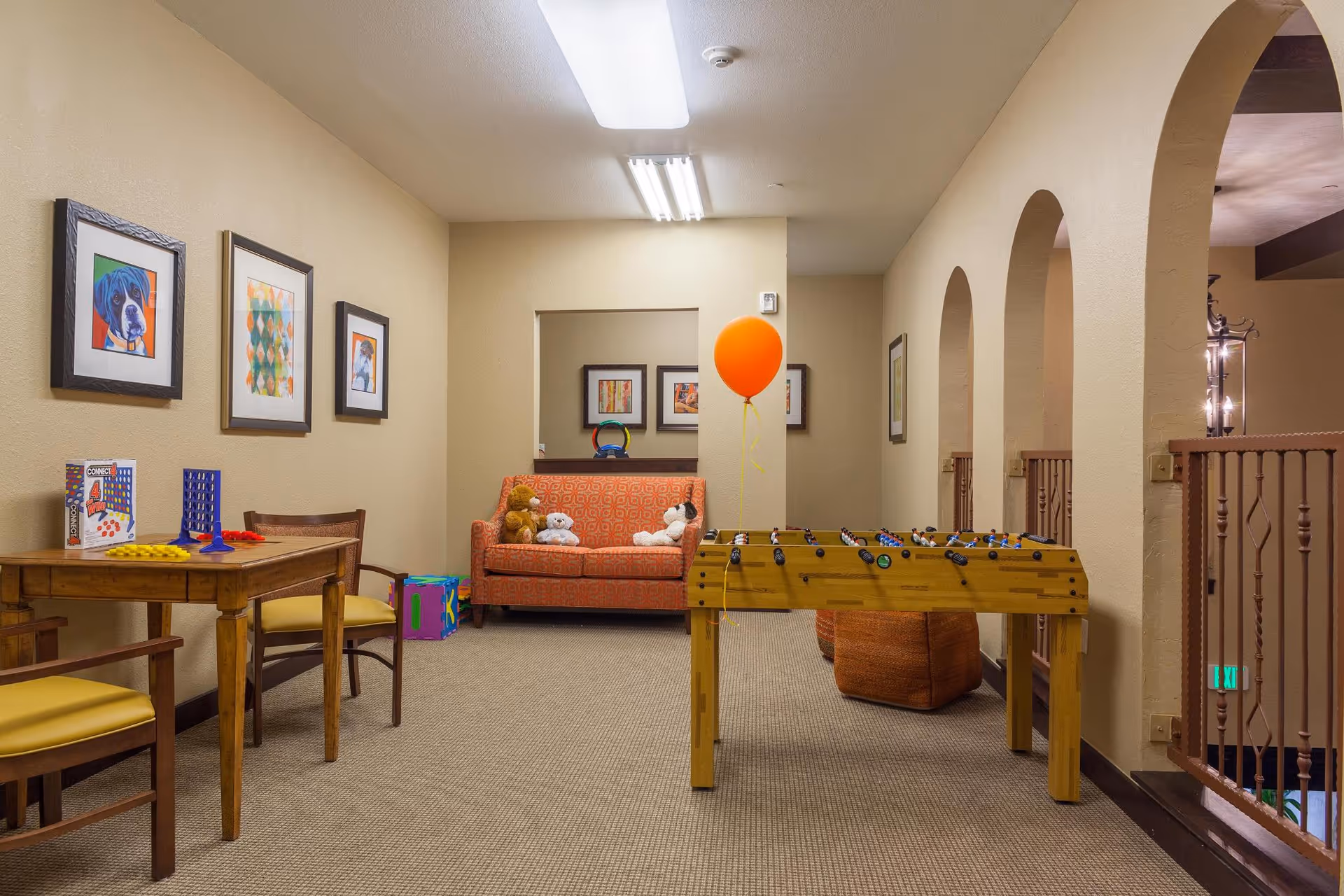 A cozy indoor recreational area with a foosball table, an orange patterned couch with stuffed animals, a wooden table with chairs and board games, and framed artwork on beige walls. There is an orange balloon tied to the foosball table and arched openings along one side of the room.