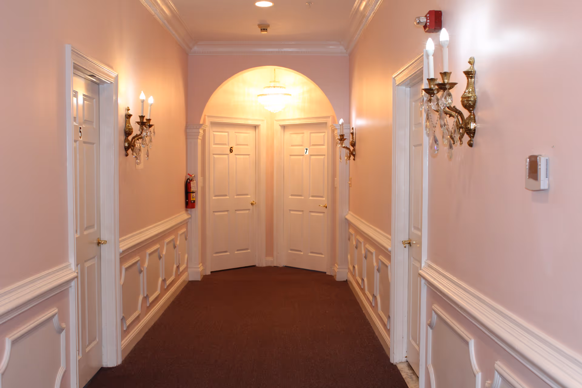 A well-lit hallway with light pink walls and white trim, featuring four white doors numbered 5, 6, and 7. The hallway has ornate wall sconces with candle-shaped bulbs and a chandelier visible through an archway at the end. A fire extinguisher is mounted on the left wall.