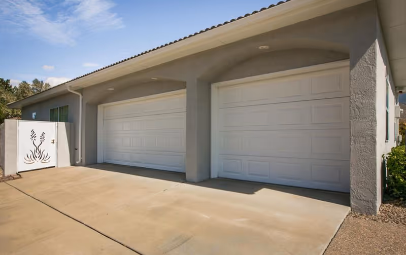 Exterior view of a building showing two white garage doors and a concrete driveway with a decorative white panel on the left.
