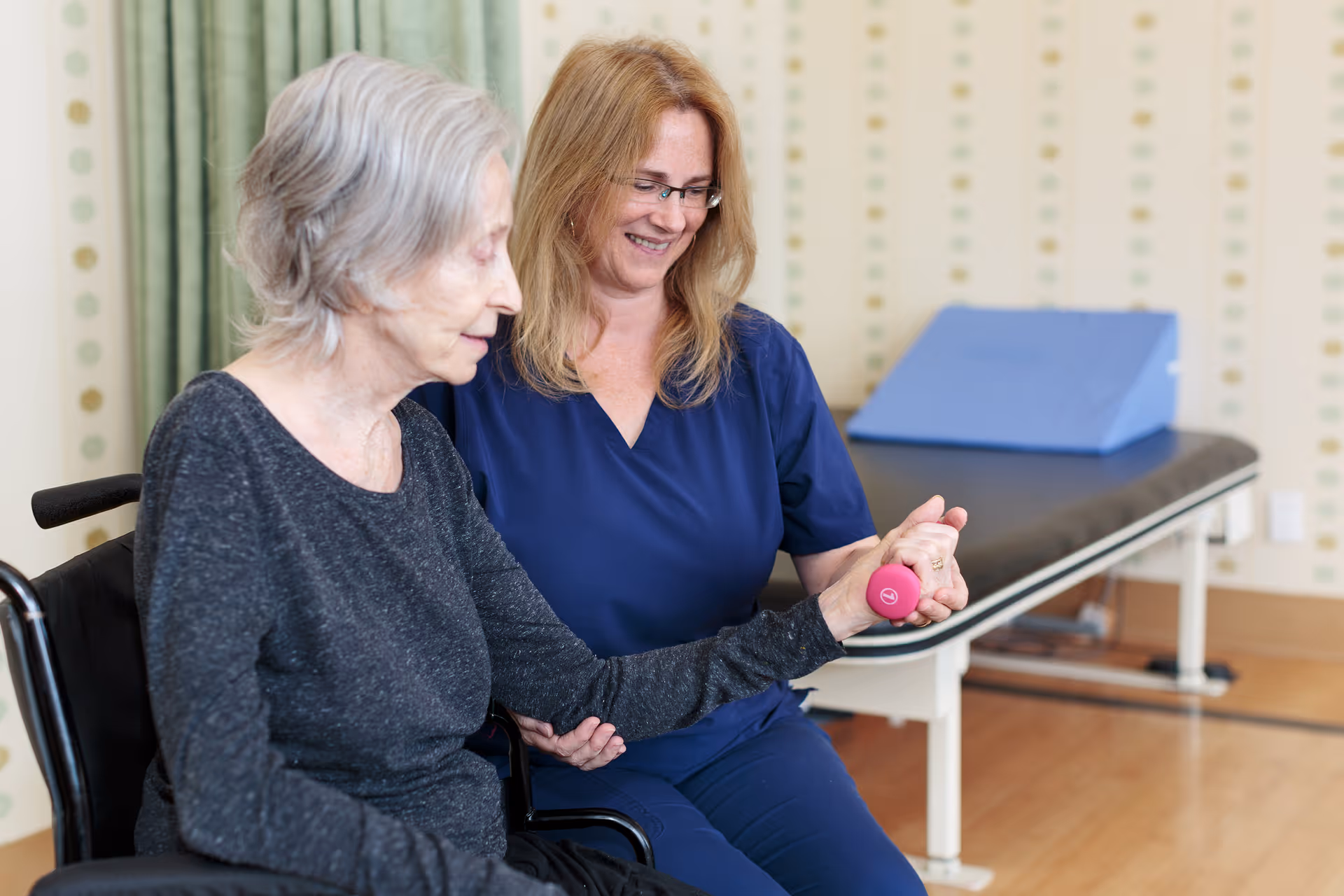 A female healthcare worker in blue scrubs assists an elderly woman in a wheelchair with a hand exercise using a small pink dumbbell in a therapy room with an examination table in the background.