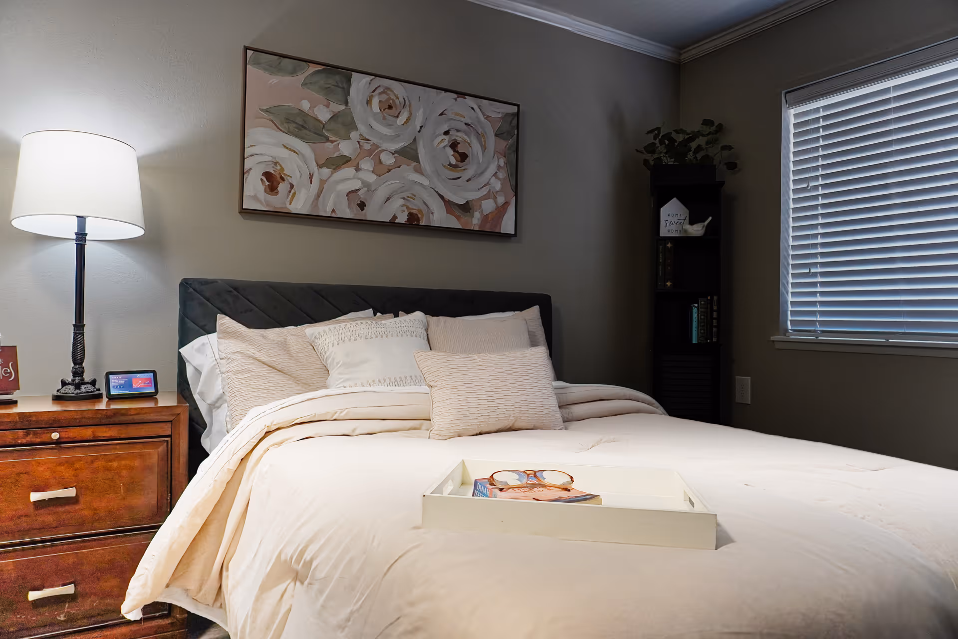 A cozy bedroom with a neatly made bed featuring multiple pillows and a white comforter. A wooden nightstand with a lamp and a small digital clock is on the left side of the bed. Above the bed hangs a floral painting with white and pink flowers. To the right, there is a window with closed blinds and a tall black bookshelf with decorative items and books.