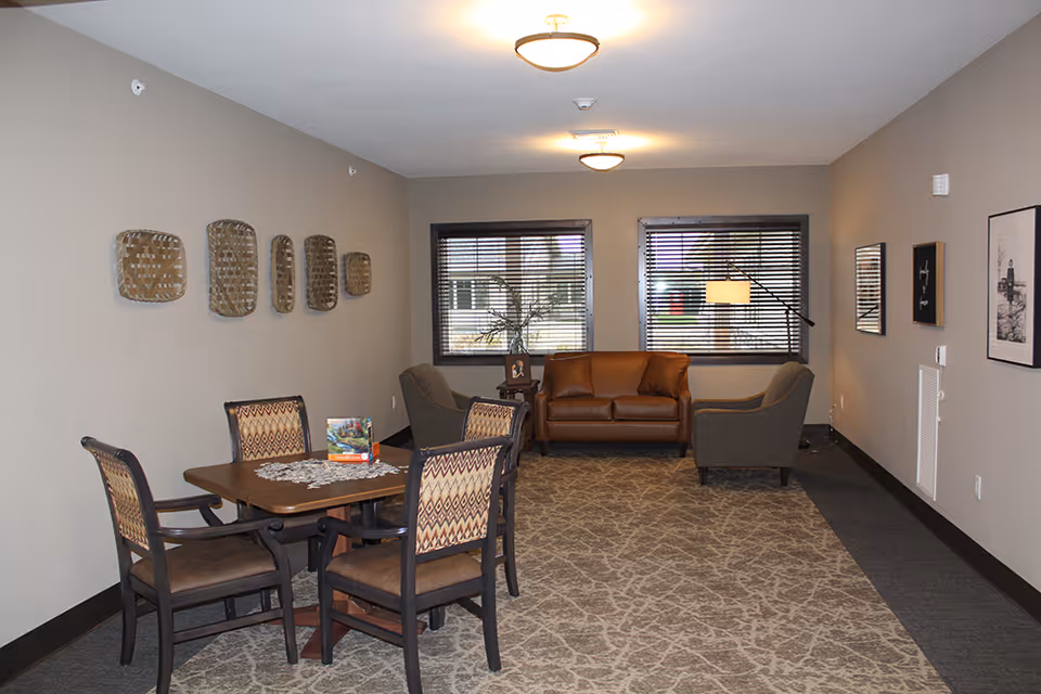 A cozy common area in The Legacy of DeForest featuring a wooden table with four patterned chairs on the left side, a brown leather loveseat, and two gray armchairs near two windows with blinds. The walls are decorated with woven baskets and framed pictures, and the floor is covered with a patterned carpet.