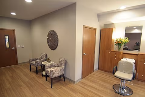 Interior view of a room with wooden flooring, two patterned armchairs with a small round table between them, a wooden door, and a salon-style chair in front of a wooden cabinet with a mirror and a vase of flowers on top.