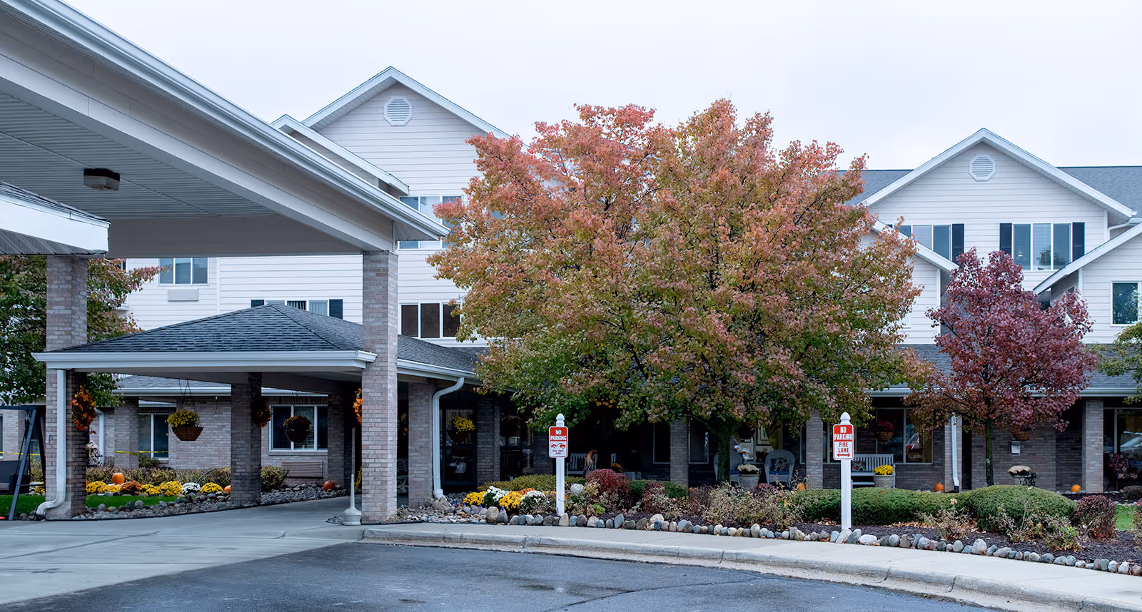 Front entrance of Holiday Genesee Gardens with a covered porte-cochère, autumn trees, and landscaped flower beds.