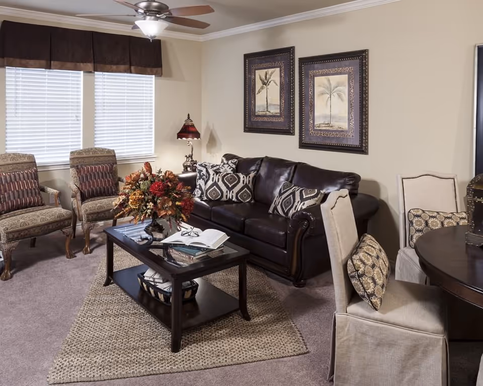 A cozy living room with a dark brown leather sofa adorned with patterned pillows, two upholstered armchairs with striped cushions, a dark wooden coffee table with a floral arrangement and books, a beige area rug, and two framed botanical prints on the beige wall. There is a ceiling fan with light, a window with blinds and a brown valance, and a round dining table with beige slipcovered chairs nearby.