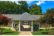 Outdoor view of a single-story building with a covered gazebo structure in the foreground, surrounded by green grass, trees, and flowering bushes under a blue sky with some clouds.