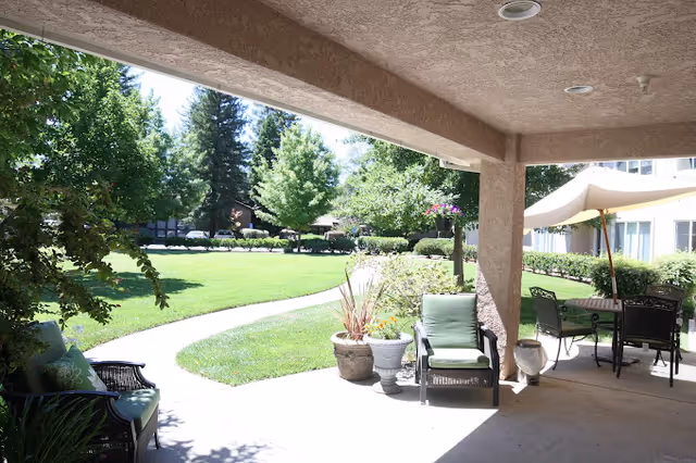 Covered patio area with outdoor seating including cushioned chairs and a table with an umbrella, overlooking a well-maintained green lawn with trees and shrubs in the background.
